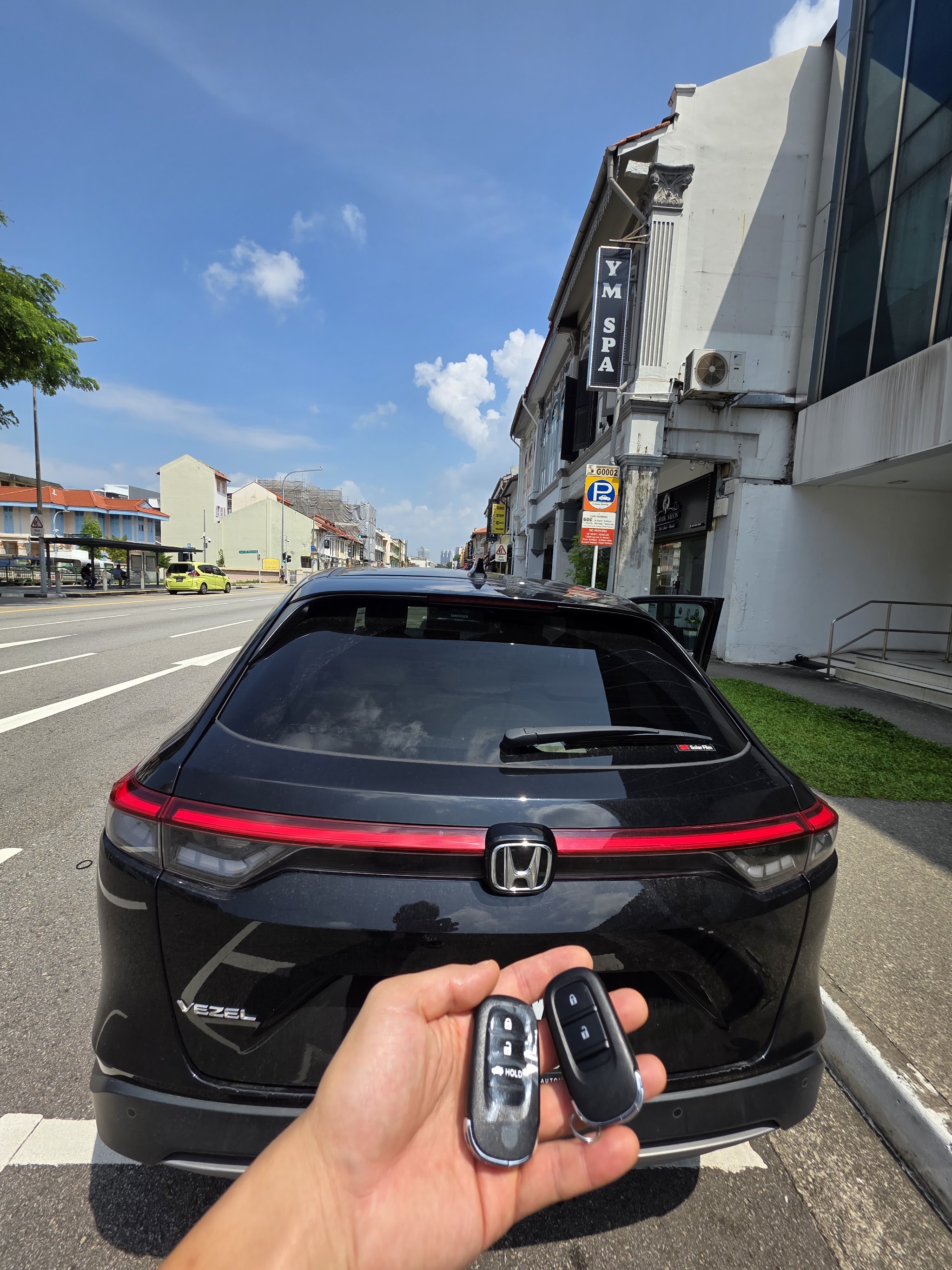 Two Honda keys held up behind a black Honda Vezel parked on Geylang Road